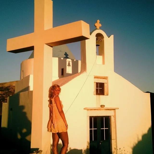 Meg Distner standing beside a tall white cross in front of a whitewashed chapel, lit by low evening sun.