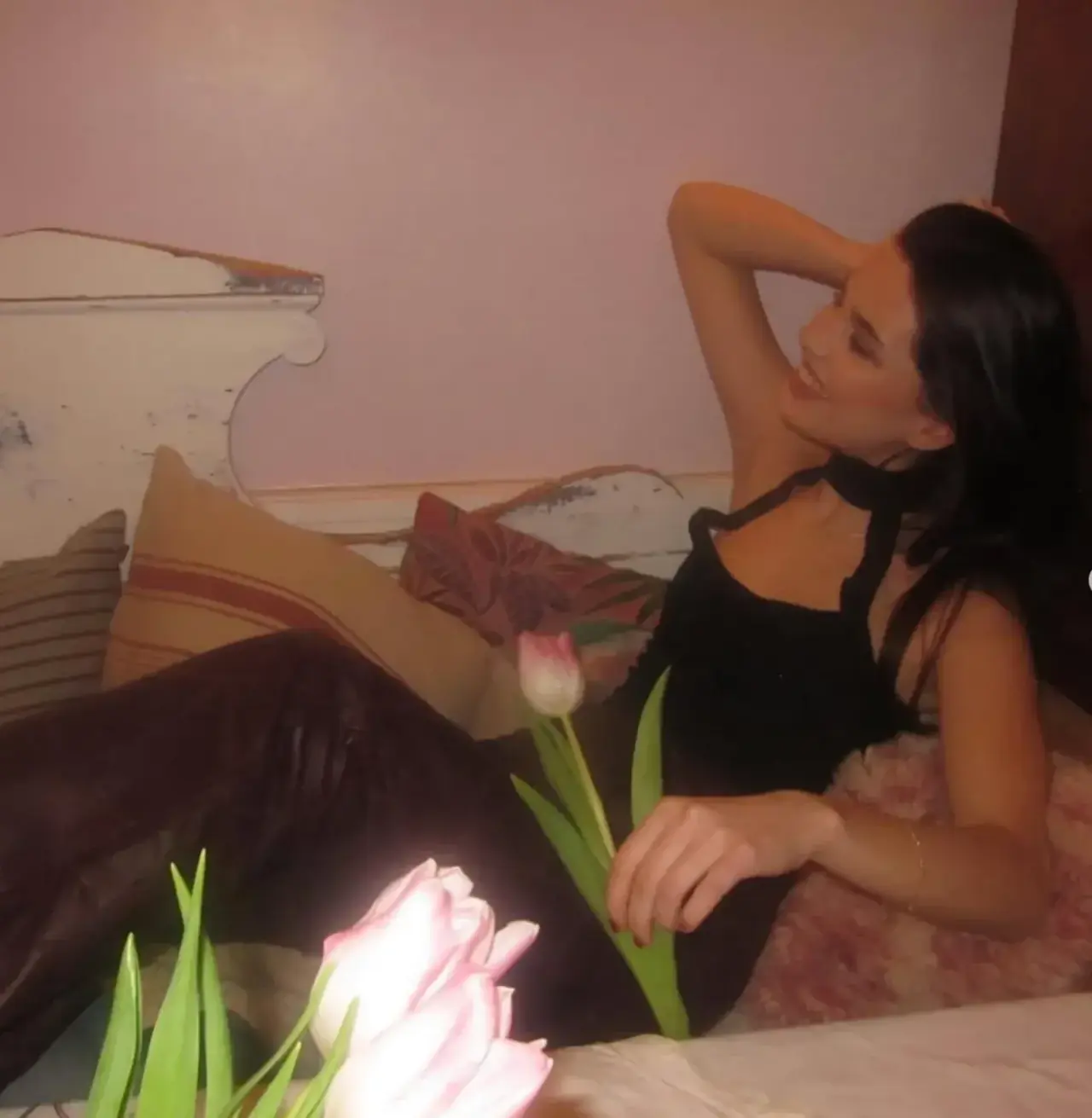 A black halter top with a neck tie worn with burgundy leather trousers, photographed reclining on a bed with tulips in the foreground.