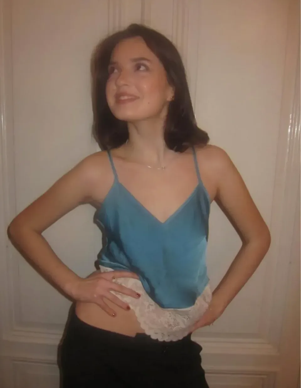 A blue satin camisole with lace trim at the hem, worn with dark trousers, hands on hips.