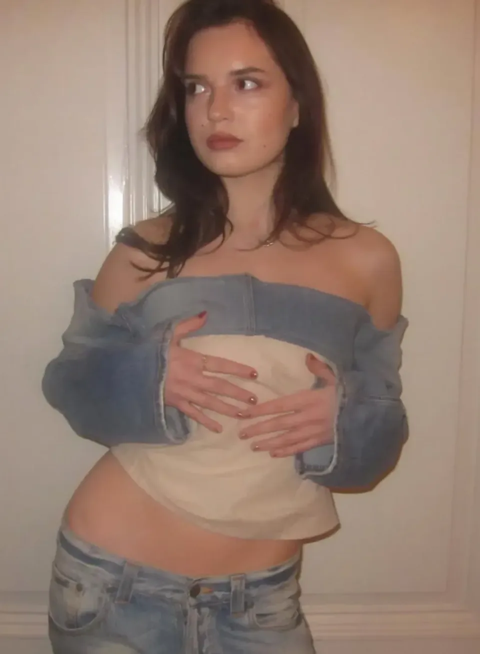 A denim jacket worn off the shoulders with a cream top held to the front, over jeans.
