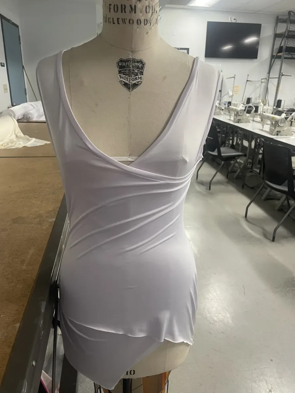 A white jersey dress draped on a dress form in the studio, with a soft cowl at the neckline and the fabric falling on the bias.