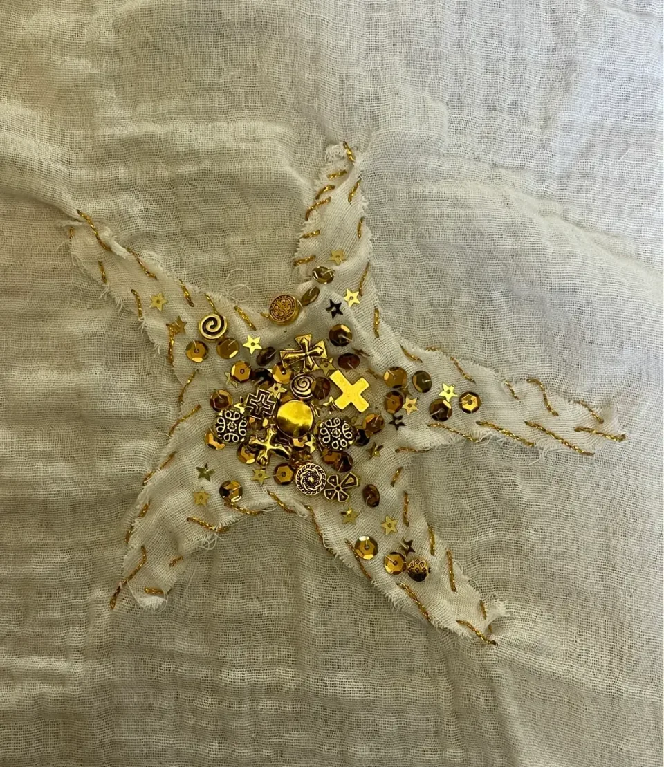 Photograph of the sewn sample: a frayed star in white double gauze, couched with gold thread and filled with sequins, beads and charms at the centre.